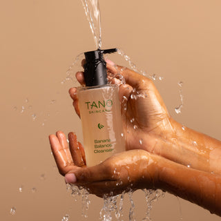 Hand holding a bottle of Tano Skincare's Banana Balance Cleanser with water being poured over it against a beige background