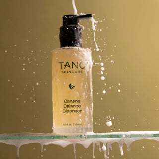 Tang Skincare Banana Balance Cleanser bottle with liquid splashing around on a gold background
