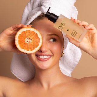 Woman with a towel on her head holding a grapefruit and Tano Skincare's Banana Balance Cleanser against a beige background